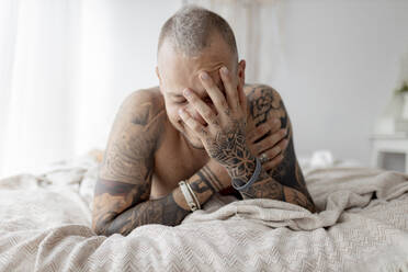 Tätowierter Hipster mit Hand im Gesicht, der zu Hause auf dem Bett liegt - GMLF00411