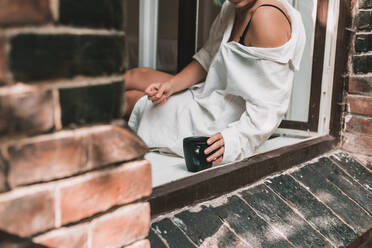 Große schwarze Kaffeetasse in den Händen einer Frau mit pastellfarbener Maniküre, die auf der Fensterbank sitzt, Lifestyle. - CAVF88035
