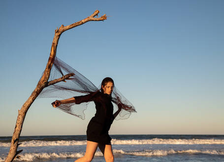 Seitenansicht einer jungen Frau, die sich in einem Fischernetz verheddert, das an einem Baumzweig an einer Sandküste mit wogendem Meer und blauem Himmel befestigt ist - ADSF09340