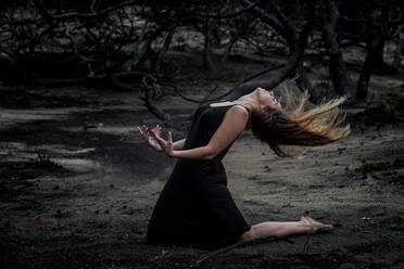 Side view of young ballerina in black wear posing on land between dry woods - ADSF09351