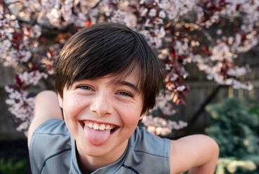 Porträt eines fröhlichen Jungen, der seine Zunge in einem Garten herausstreckt. - CAVF88059