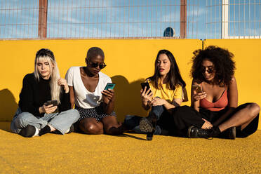 Careless youthful diverse women in casual clothes messaging on cellphone while sitting on asphalt ground in stadium - ADSF09375