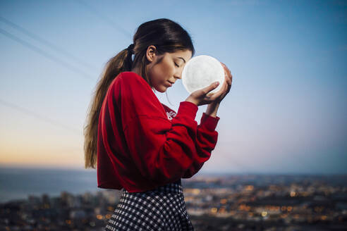 Young woman with eyes closed holding moon shape lamp against sky at dusk - MIMFF00154