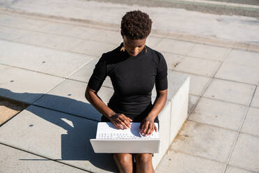 Konzentrierte afroamerikanische Frau im eleganten schwarzen Kleid, die einen Laptop benutzt, während sie sich auf dem Bürgersteig auf der Straße ausruht - ADSF09393