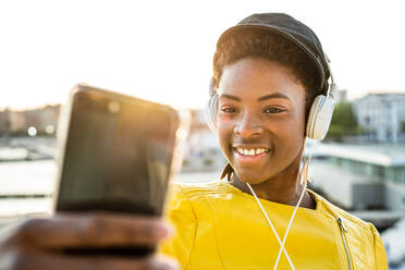 Afroamerikanische Frau in modischer heller Jacke, die ein Mobiltelefon benutzt und über Kopfhörer Musik hört - ADSF09400