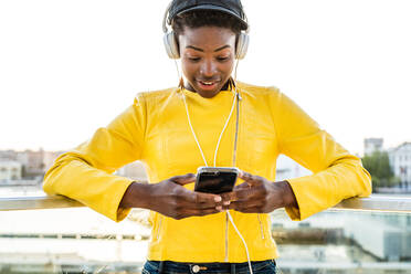 Afroamerikanische Frau in modischer heller Jacke, die ein Mobiltelefon benutzt und über Kopfhörer Musik hört - ADSF09401