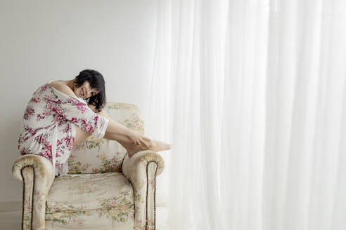 Frau in Nachtwäsche sitzt zu Hause auf einem Sessel - GMLF00414