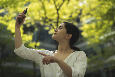 Junge Frau nimmt Selfie durch Smartphone gegen Bäume - HPSF00055