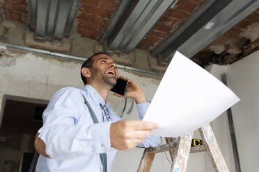 Lachender Architekt mit Plan am Telefon in einem im Bau befindlichen Haus - VABF03305