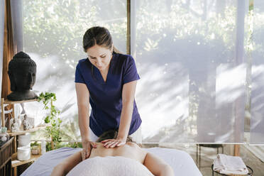 Mid adult woman giving back massage to female customer lying on table in spa - EBBF00512