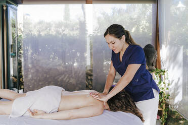 Smiling female therapist giving back massage to woman lying on table in spa - EBBF00515