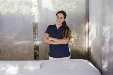 Lächelnde weibliche Massagetherapeutin mit verschränkten Armen, die an einem Tisch im Spa steht - EBBF00540