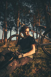 Nachdenklicher junger Mann sitzt bei Sonnenuntergang mit dem Fahrrad im Park - ACPF00785