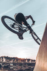 Carefree young man performing stunt with bicycle on ramp against sky in park during sunset - ACPF00789