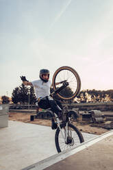 Carefree man wearing helmet performing stunt with bicycle on ramp in park at sunset - ACPF00792