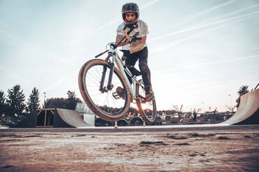 Young man wearing helmet riding bicycle on ramp in park at sunset - ACPF00793