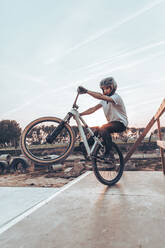 Young man wearing helmet riding bicycle on ramp in park against sky during sunset - ACPF00796
