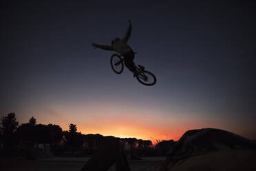 Carefree silhouette man performing stunt with bicycle against clear sky during sunset - ACPF00798