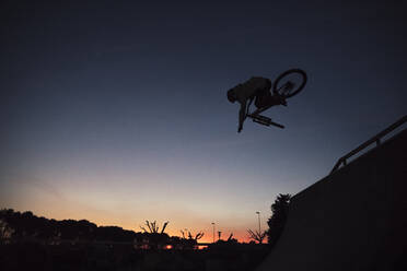 Silhouette young man performing stunt with bicycle against clear sky during sunset - ACPF00799