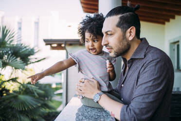 Father holding baby girl pointing on retaining wall while standing in balcony - MEUF01880