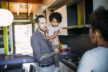 Vater trägt seine Tochter bei einem Gespräch mit einer Frau in der Küche zu Hause - MEUF01892