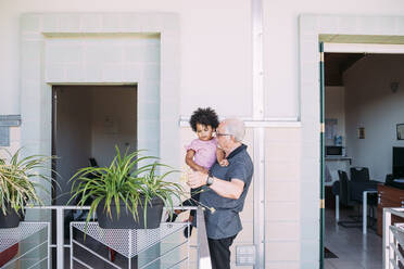 Grandfather carrying baby granddaughter while standing in balcony - MEUF01903