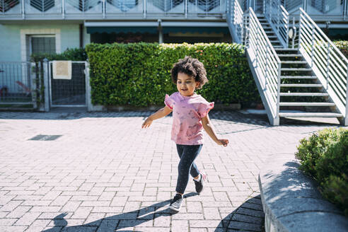 Baby-Mädchen mit lockigem Haar zu Fuß auf dem Gehweg während sonnigen Tag - MEUF01906