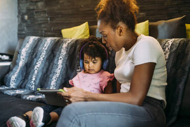 Mutter zeigt ihrer Tochter mit Kopfhörern ein digitales Tablet, während sie zu Hause auf dem Sofa sitzt - MEUF01915