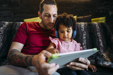 Vater mit Tochter, die auf dem Sofa zu Hause ein digitales Tablet benutzt - MEUF01917