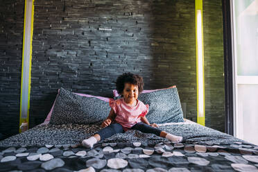 Smiling baby girl with legs apart sitting on bed against wall at home - MEUF01918