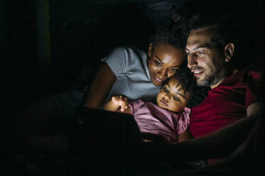 Lächelnde Eltern mit Tochter, die ein digitales Tablet benutzt, während sie sich zu Hause auf dem Bett entspannen - MEUF01921