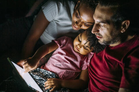 Eltern mit Tochter nutzen ein digitales Tablet, während sie sich zu Hause im Bett entspannen, lizenzfreies Stockfoto