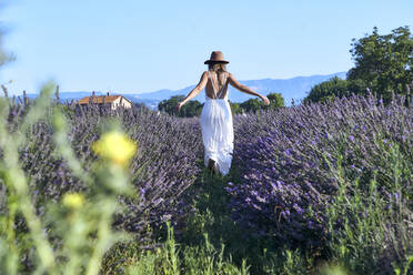 Woman wearing white dress and hat walking amidst lavender field against clear sky - VEGF02616