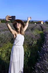 Smiling woman wearing white dress taking selfie with smart phone while standing amidst lavender field - VEGF02624