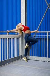 Young woman dancing by metal railing in front of blue wall in the city - MAUF03514