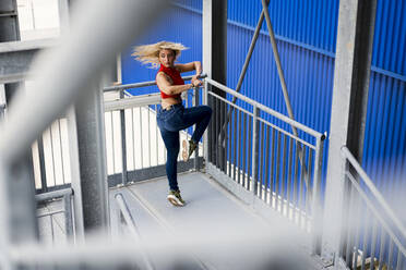 Junge Frau tanzt an einem Metallgeländer vor einer blauen Wand in der Stadt - MAUF03515