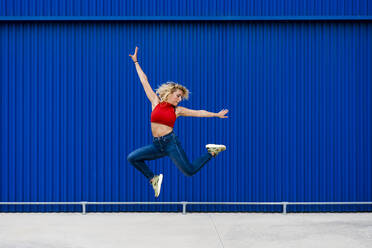 Junge Frau springt vor einer blauen Wand in der Stadt - MAUF03517
