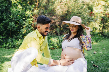 Glücklicher Mann, der den Bauch einer schwangeren Frau berührt, während er im Park sitzt - DCRF00583