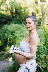 Schwangere Frau hält Babyschuhe, während sie in einem öffentlichen Park steht - DCRF00601