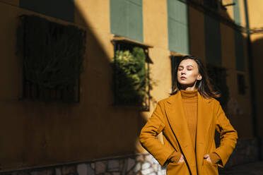 Young woman in winter coat standing with eyes by building - TCEF00967