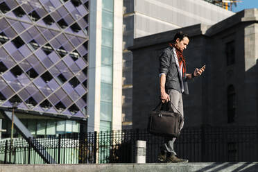 Trendy Mann mit Handtasche mit Smartphone beim Spaziergang in der Stadt - JMPF00291