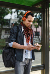Trendiger Mann, der sein Smartphone benutzt, während er an der Bushaltestelle über Kopfhörer Musik hört - JMPF00295