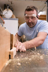 Low angle of focused carpenter in protective glasses milling timber while crafting detail using electric machine in light modern workshop - ADSF09421