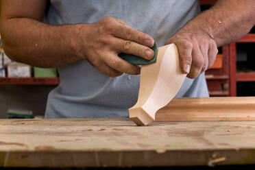 Niedriger Winkel eines gesichtslosen Schreiners, der ein Holzteil mit Schleifpapier poliert, während er in einem hellen, modernen Arbeitsraum arbeitet - ADSF09422