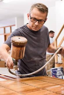 Handwerker beim Auftragen von Lack auf eine Holzjalousie mit Airbrush in einer Tischlerei - ADSF09427