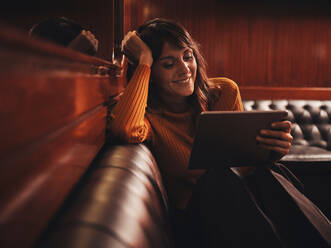 Stylish smart glückliche Frau Surfen Tablet bequem sitzen mit den Beinen auf schwarzem Ledersofa in Cafe - ADSF09431