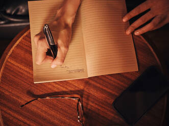 From above crop unrecognizable woman writing with pen in notebook at round wooden table with mobile phone and glasses in cafe - ADSF09434