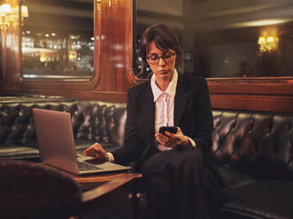 Smart business woman in glasses typing on laptop and checking information in mobile phone comfortably sitting in black leather sofa in cafe - ADSF09441