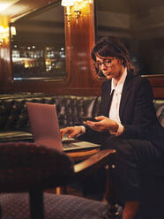 Smart business woman in glasses typing on laptop and checking information in mobile phone comfortably sitting in black leather sofa in cafe - ADSF09442