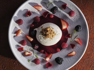 Walnut grated on creamy dessert with jam surrounded by pieces of strawberry blueberry and raspberry on white dish - ADSF09481